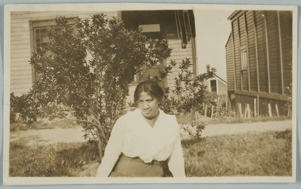 A black-and-white snapshot photograph of a Black woman posing in front of a shrub blocking a house in the background.