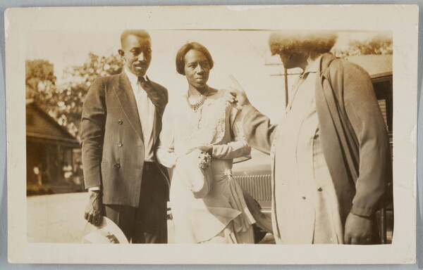 A black-and-white snapshot photograph of a Black man and woman standing next to what looks like a car; a third person is turned to them pointing.