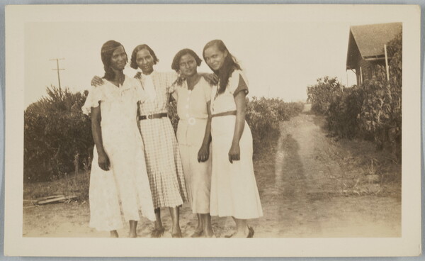 A black-and-white snapshot photograph of four Black women, all wearing dresses, standing with their arms around each other; the photographer's shadow is on the grass next to them.