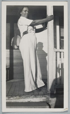 A black-and-white snapshot of a Black woman in white shirt and pants standing on a porch holding onto a column.