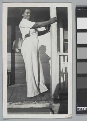 A black-and-white snapshot of a Black woman in white shirt and pants standing on a porch holding onto a column.