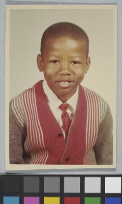 A color school photograph of a young, smiling Black boy wearing a white collared shirt and red tie under a striped cardigan.