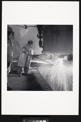 A black-and-white photograph of an industrial worker, wearing safety helmet, coat, and gloves holding a rod under a machine emitting sparks.