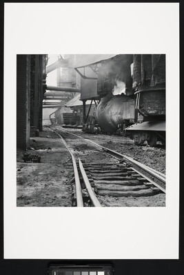 A black-and-white photograph of railroad track  between industrial buildings and  machinery, some emitting steam or smoke.