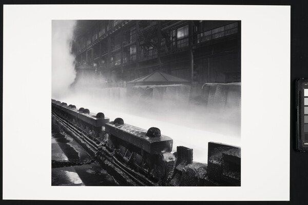 A black-and-white photograph of industrial machinery in front of steam or smoke rising near a multi-story industrial building.