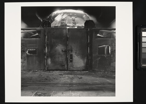 A black-and-white photograph of a metal wall with large doors behind which is an industrial furnace.