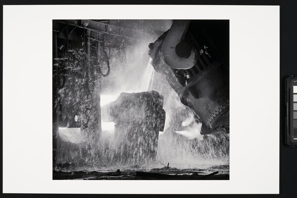 A black-and-white photograph of a piece of machinery pouring molten metal into another piece of machinery, all surrounded by sparks.