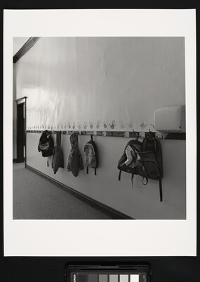 A black-and-white photograph of a hallway with kids' backpacks and jackets hanging on hooks.