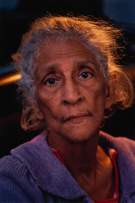 A color portrait photograph of an older Black woman with white hair and sunken cheeks, wearing a lavender sweater.