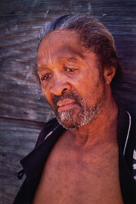 A color portrait photograph of a Black man, salt-and-pepper hair slicked back, gray beard, downward gaze, wearing a black jacket open to reveal his bare chest.