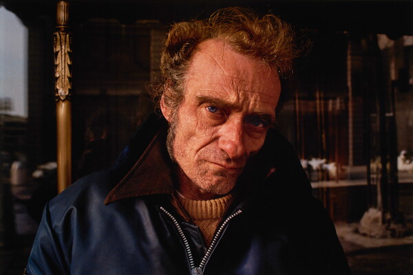 A color portrait photograph of a White man with a wrinkled face, thick brows, long sideburns, and sunken cheeks wearing a blue jacket with a tan collar.