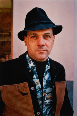 A color portrait photograph of a fair-skinned man wearing a fedora hat and a blue Hawaiian print shirt under and black and brown jacket.