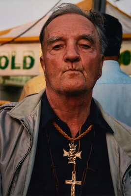 A color portrait photograph of a medium-skinned man with gray hair, lips pursed, wearing several gold cross necklaces over a black shirt.
