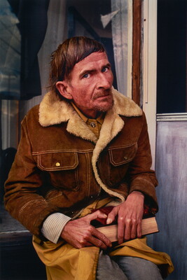 A color portrait photograph of a seated middle-aged White man, short hair, a stubbly goatee, wearing a brown coat lined with cream fleece, holding a book in his lap.