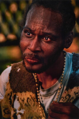 A color portrait photograph of a Black man with a goatee wearing a beaded necklace, and a torn cutoff vest over a t-shirt.