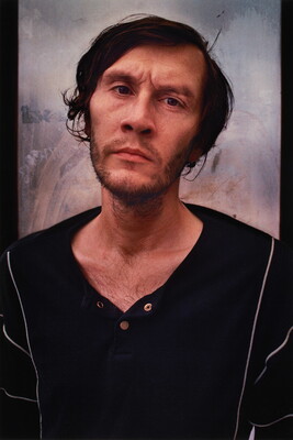A color portrait photograph of a White man with short untidy hair, a stubbly beard and mustache, a furrowed brow, wearing a black open-necked shirt.