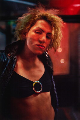 A color photograph of a young White person with slightly matted hair wearing a jacket over a bikini top; red lights in the background reflect on their face.