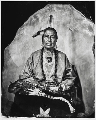 A black-and-white portrait photograph of a seated Indigenous person, feathers in their hair, strings of beads around their neck, holding several objects in their hands.