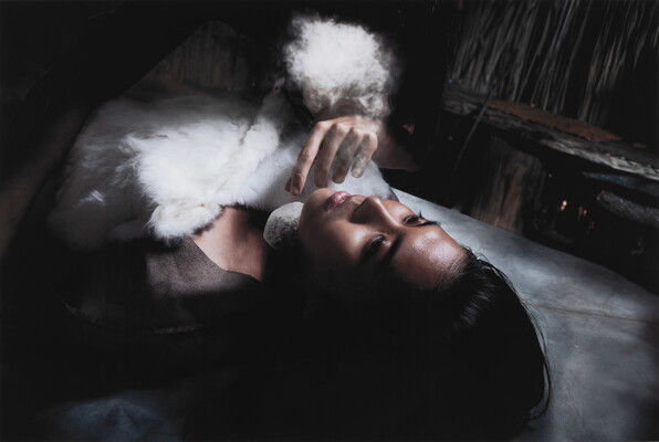 A color photograph of a young Indigenous woman lying on her back, eyes open, chest obscured by fluffy cloudlike shapes, as a pale hand reaches toward her face.