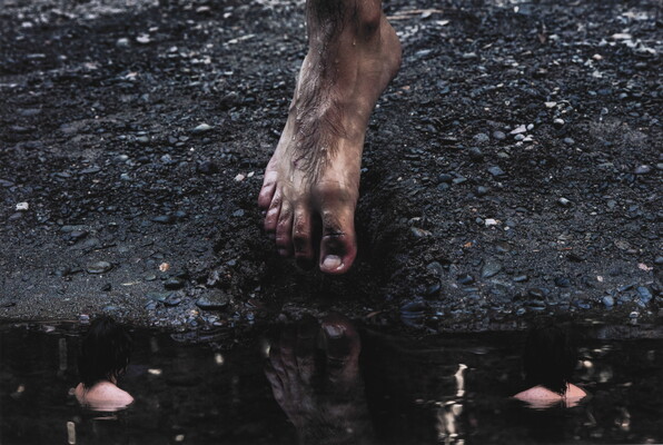 A color photograph of a masculine foot lifting off a muddy, gravelly bank about to step into the dark water in which it is reflected.