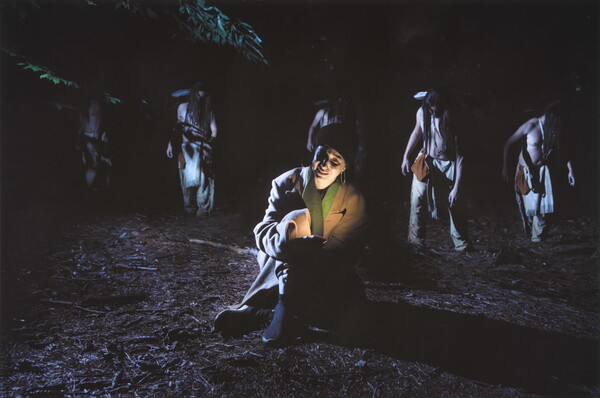 A color nighttime photograph of five Indigenous figures behind a White woman seated on the ground holding a smartphone that lights her face in the darkness.