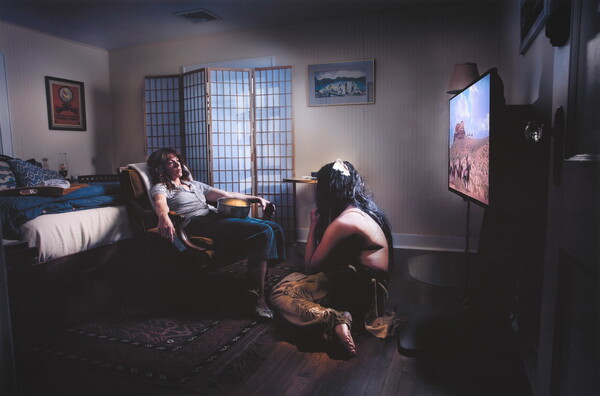 A color photograph of a White woman lying back in a chair, TV remote in one hand and a bowl of popcorn on her lap, as a Native American man sits on the floor between her and a television.