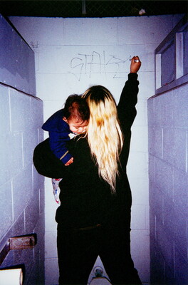 A color photograph of a woman, her back to the viewer, holding a baby while writing graffiti on a bathroom stall wall.