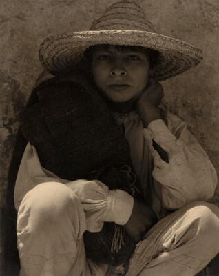 A black-and-white photograph of a young medium-skinned boy wearing a straw hat, crouched in front of a wall.