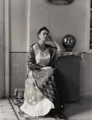 A black-and-white photograph of a seated, young, medium-skinned woman, hair pulled up, wearing a shawl over her long dress; one elbow rests on the table next to her on which sits a reflective globe.