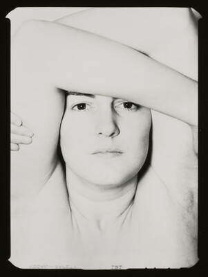 A black-and-white photograph of the face of a young White woman with dark eyes, wrapping her arms up over her head.