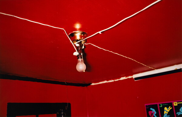 A color photograph of the ceiling of a red-painted room with exposed wires and a bare lightbulb hanging from the ceiling.