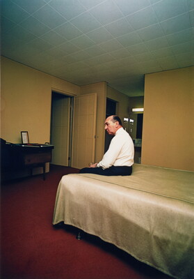 A color photograph of an older, balding White man sitting on a bed in a sparsely furnished room.