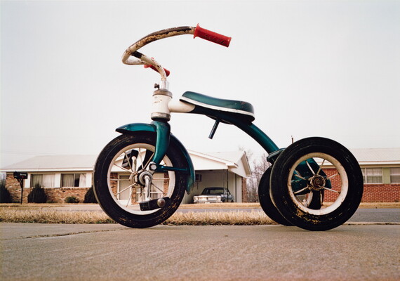 A color photograph using perspective to make a rusty green tricycle in the foreground appear larger than the houses in the background.