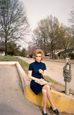 A color photograph of a White woman with styled blond hair in a dark blue dress sitting on a neighborhood street curb.