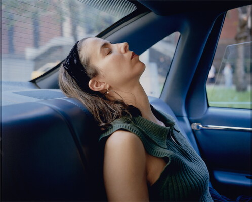 A color photograph of a White woman, eyes closed, black headband in her hair, leaning her head back on the backseat of a car.