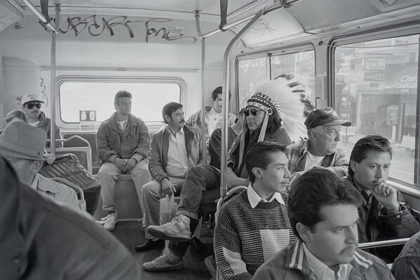 A black-and-white photograph of an Indigenous man wearing a full feathered headdress sitting on a crowded public bus.