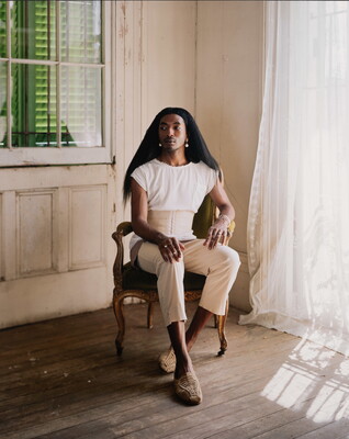 A color photograph of a young Black man with long hair, wearing a white shirt and tan pants, seated in a room next to a window with flowing white curtains.