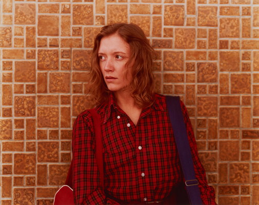 A color photograph of a White woman with reddish hair wearing a plaid, button-down shirt standing against a tile wall and looking to her right.