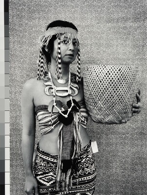 A black-and-white photograph of a medium-skinned woman wearing a headband with long beaded fringes, a halter top, patterned skirt, and holding a woven basket.