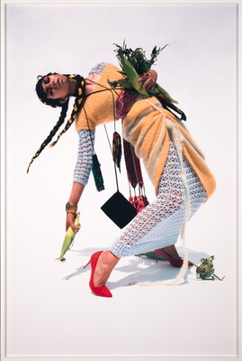 A color photograph of a young Indigenous woman, hair in braids, wearing an orange wrap over a white dress, clutching ears of corn in one arm and holding one ear in her other hand, leaning back as if falling.