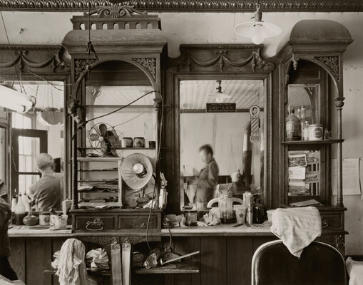 A black-and-white photograph of ornate shelves and counters in a barbershop cluttered with tools, and large mirrors that reflect the people in the shop.