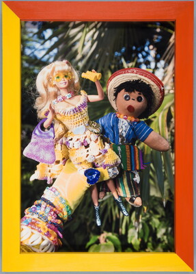 A color photograph of a hand in a yellow glove holding a Barbie wearing a yellow dress and a plush doll dressed in traditional Mexican clothing.