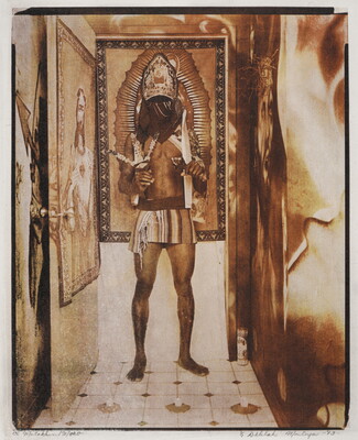 A sepia-toned photograph of a medium-skinned man in a doorway with his face covered, wearing a miter-type hat and a loincloth, and holding feathers in one hand and a mace in the other.