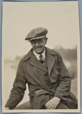 A black-and-white photograph of a smiling White man seated outside, wearing a newsboy cap, round wire-rimmed glasses, and a trench coat.