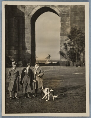 A black-and-white photograph three people and a spotted dog standing in front of a very tall stone archway.