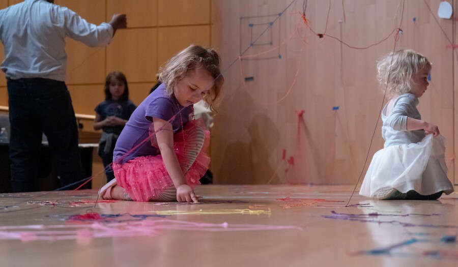 Two toddlers play with colored string on the floor of a gallery.