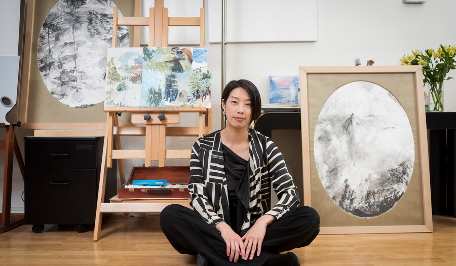 Artist Lingchia Tsai sits on the floor of her studio in front of art and easels.