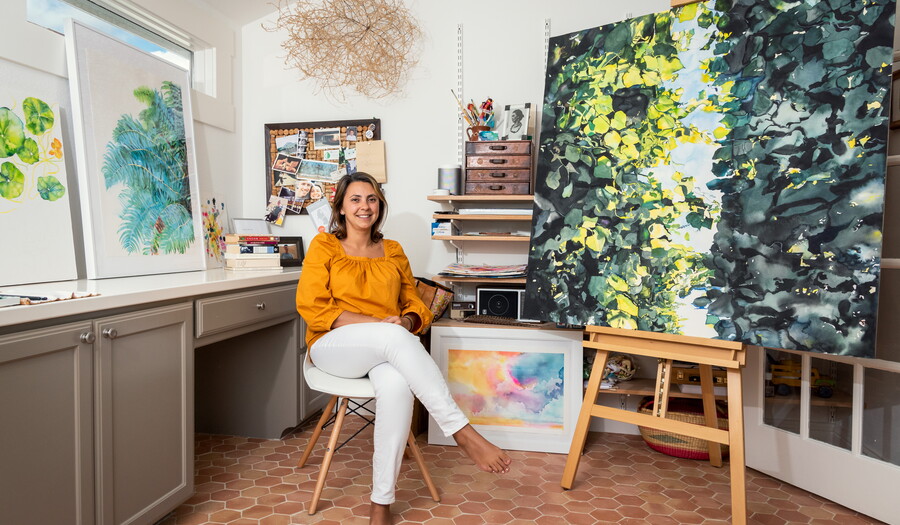 Artist Brenda Ciardiello sits in her studio surrounded by paintings on the wall and on easels.