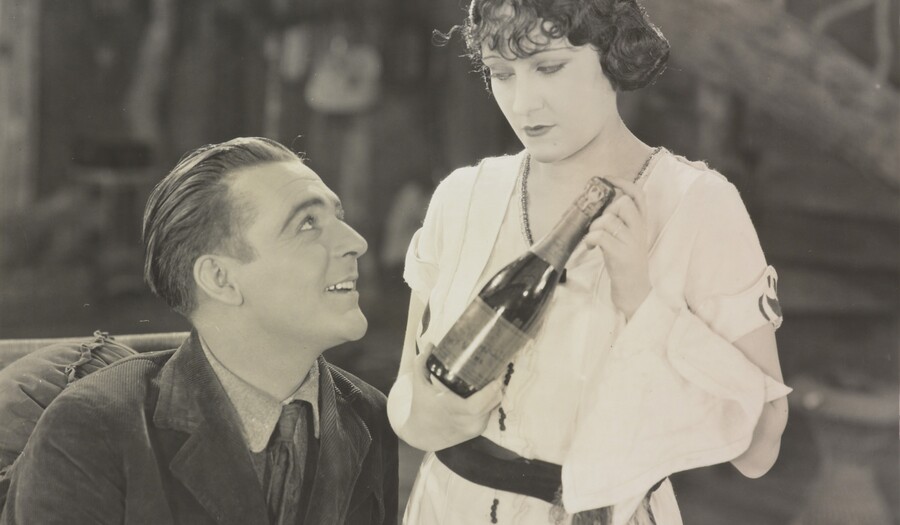 Black and white photo of a seated man looking up at a woman standing next to him holding a bottle of wine