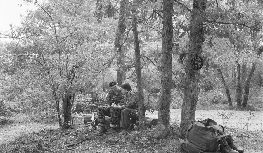 A black-and-white photograph of two people, on dressed in fatigues, sitting on a log in a wooded setting.
